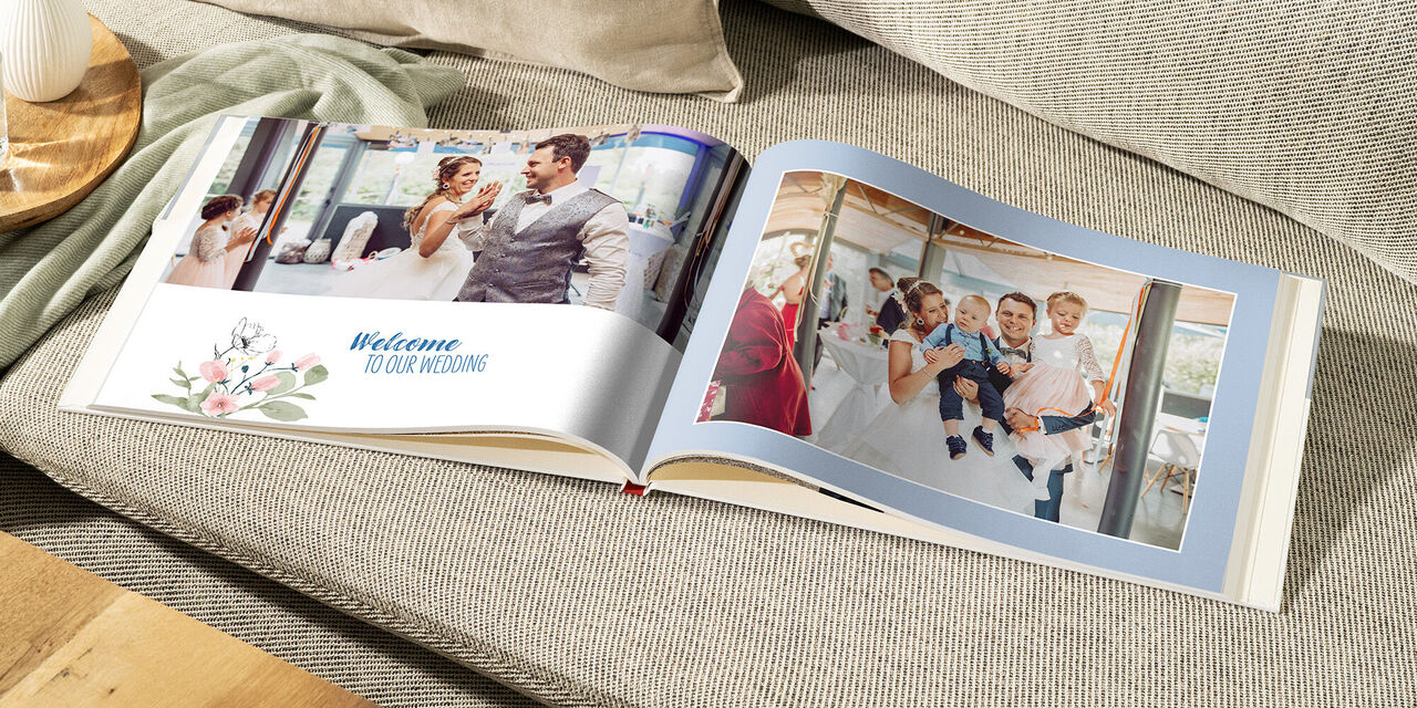 Auf einem hellen Sofa liegt ein aufgeschlagenes CEWE FOTOBUCH. Auf der linken Seite steht «Welcome to our Wedding» und man sieht das Brautpaar tanzen. Auf der rechten Seite sieht man das Brautpaar mit ihren Kindern.