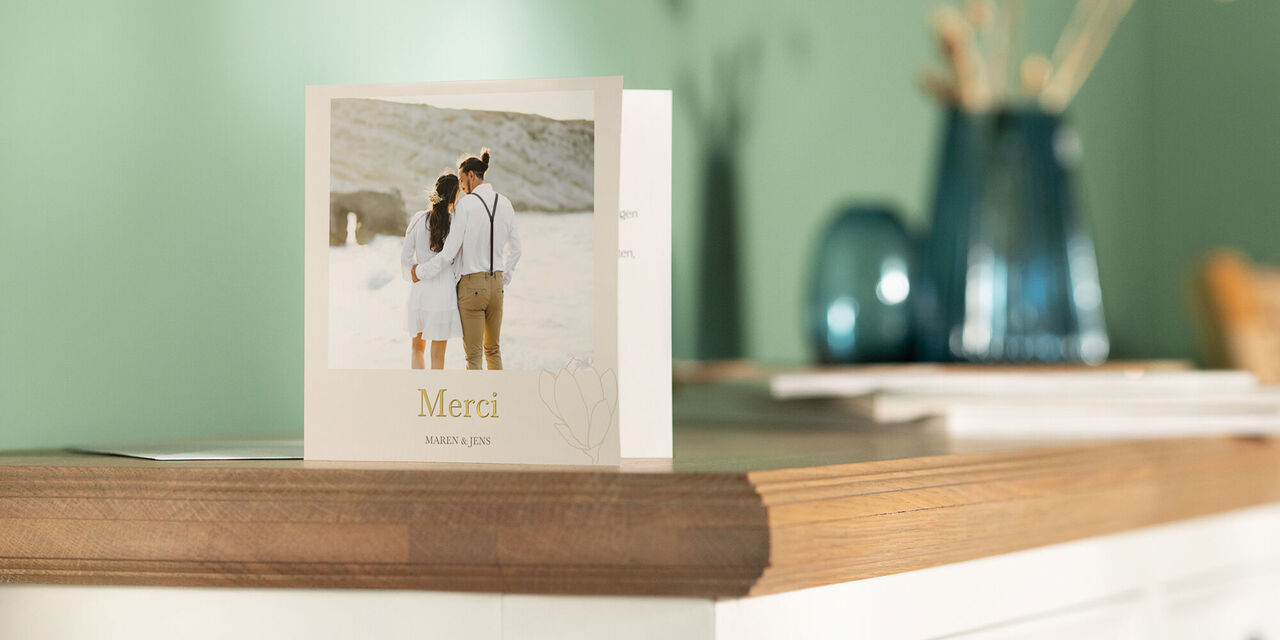 Une carte de remerciement est posée sur une commode. Au recto, on peut voir un dessin gris autour d’une photo des mariés. Sous l’image, on peut lire « Merci » en lettres dorées.